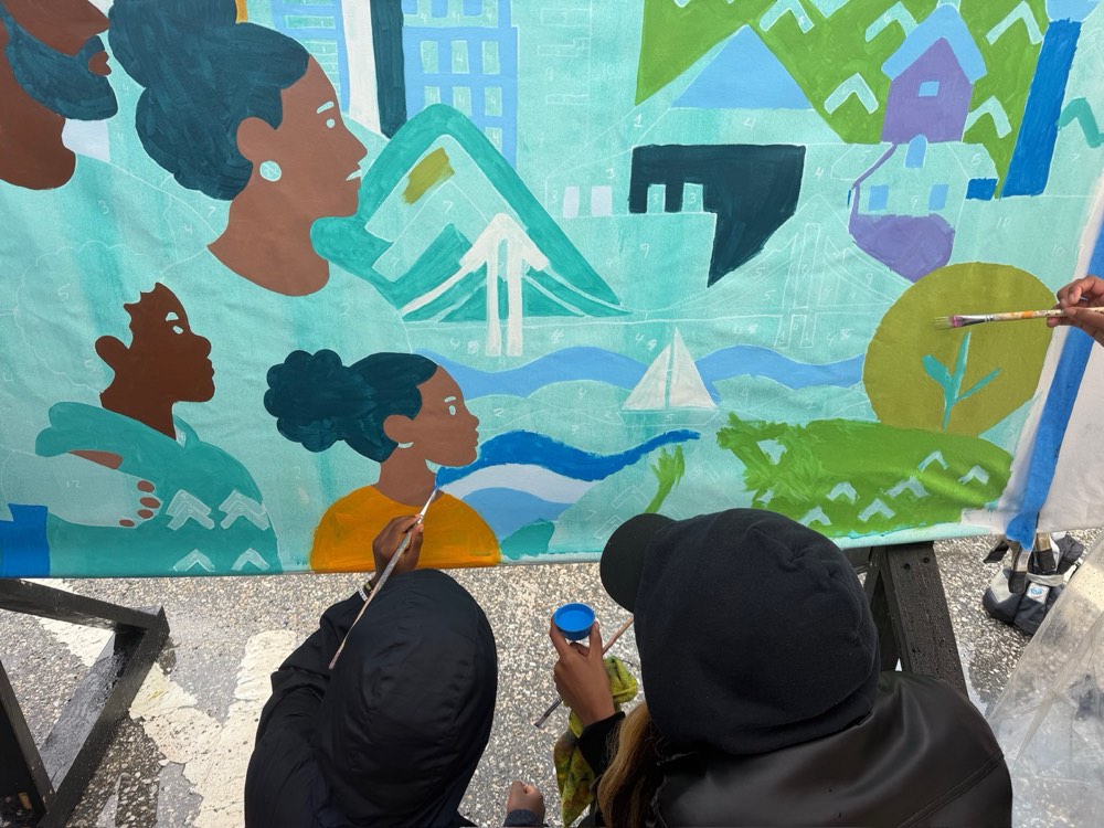 Close view of two painters adding colour to the mural's rowhouses and hills