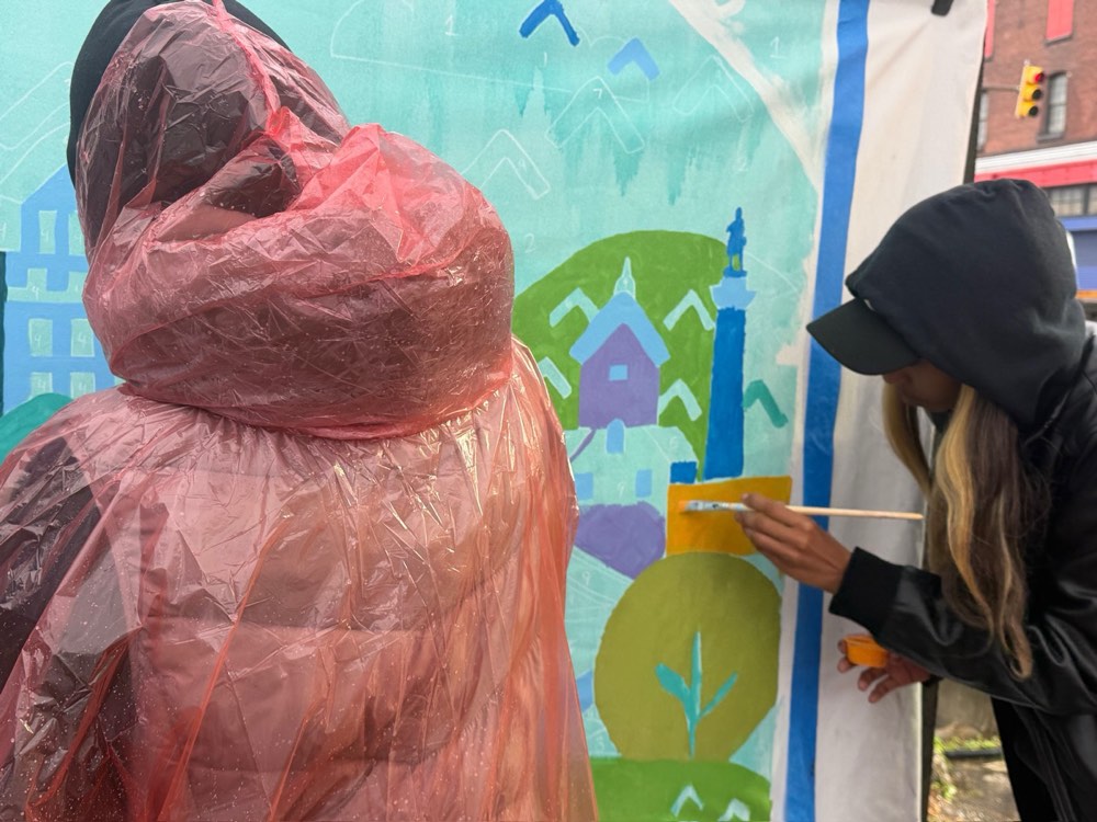 Two volunteers in rain ponchos keep painting the mural in the rain
